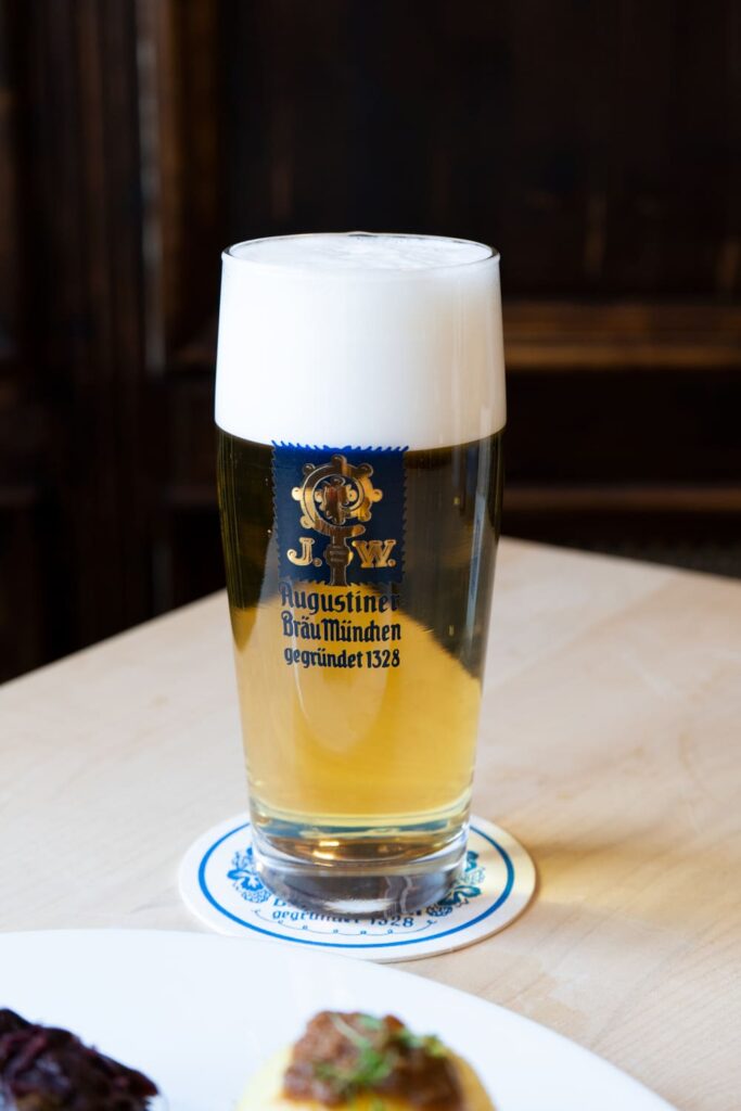Ein frisch gezapftes, helles Augustiner-Bier mit üppiger Schaumkrone auf einem Bierdeckel, serviert auf einem hellen Holztisch.