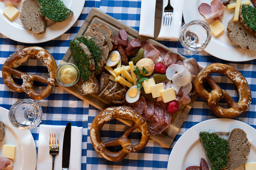 Eine bayerische Brotzeit mit Brezen, Wurst, Käse und Radieschen auf einer blau-weiß karierten Tischdecke in der Draufsicht.