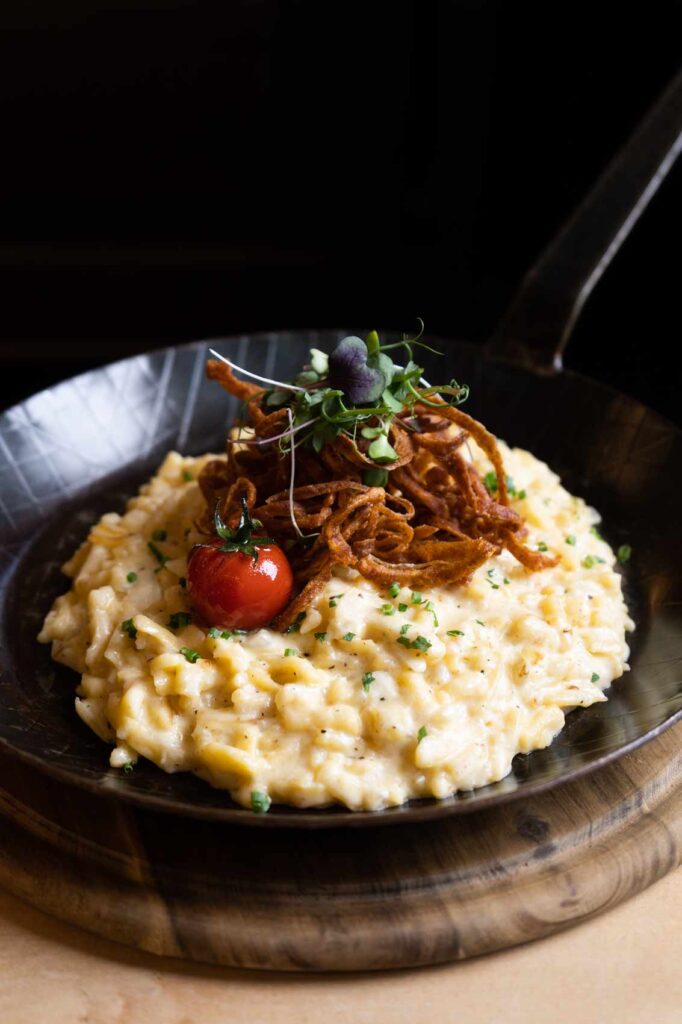 Käsespätzle in einer gusseisernen Pfanne auf einem Holzbrett, garniert mit knusprigen Röstzwiebeln, einer Kirschtomate und frischen Kräutern.