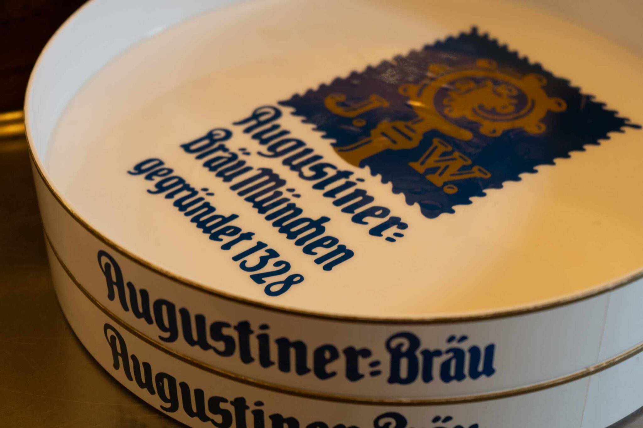 Nahaufnahme eines runden Augustiner-Bräu-Schriftzugs mit dem blauen Aschenbechers mit Logo und dem Text „gegründet 1328“.