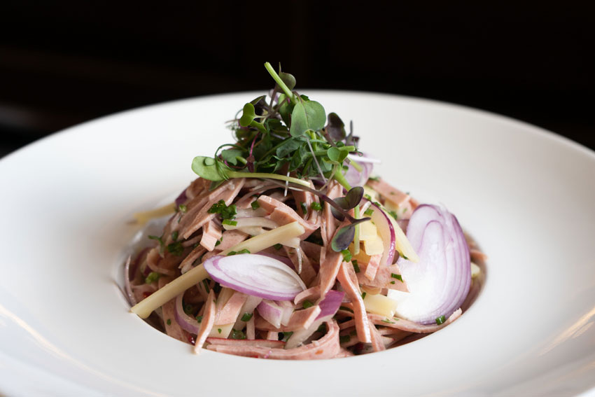 Elegant angerichteter Wurstsalat mit feinen roten Zwiebelringen und frischen Kräutern auf einem tiefen weißen Teller.