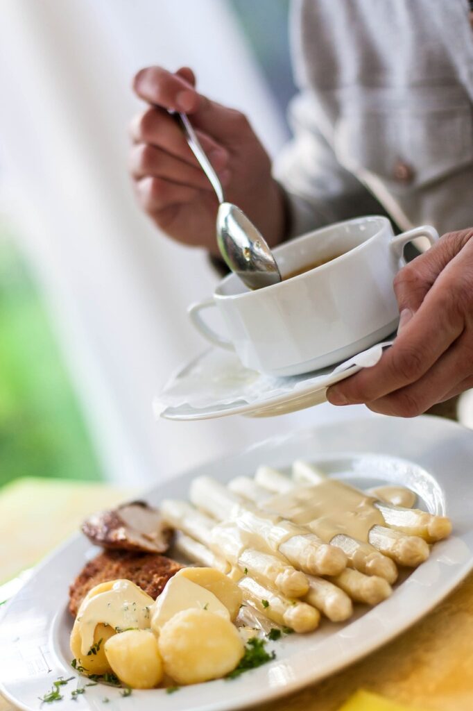 Ein Teller mit weißem Spargel, Sauce Hollandaise, Kartoffeln und Fleisch, während im Hintergrund eine Person eine Suppentasse hält und mit einem Löffel darin rührt.